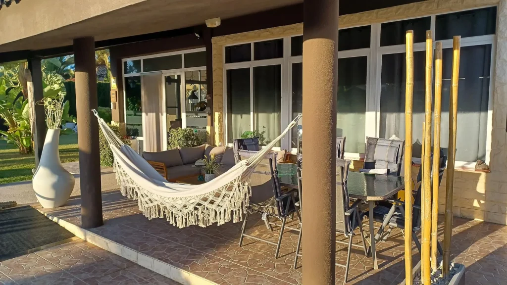 Terraza con hamaca de macramé blanca, muebles de exterior y decoración moderna en un entorno soleado.