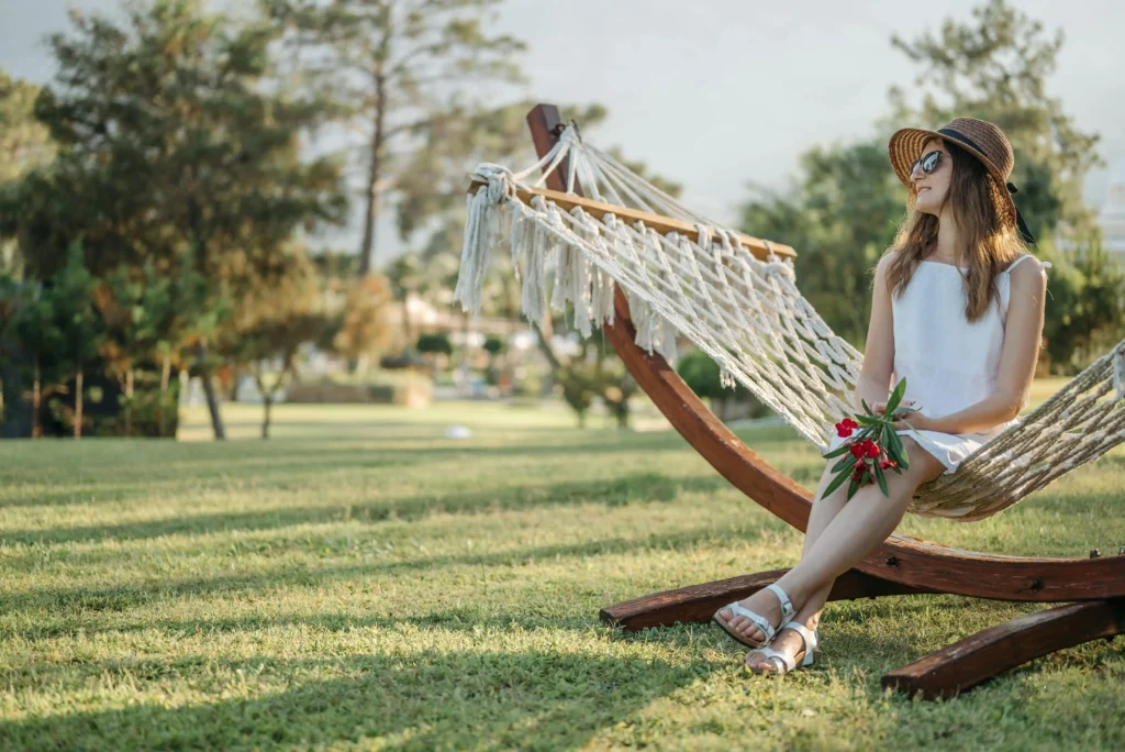 Mujer relajándose en una hamaca de cuerda en un jardín, con sombrero y flores en las manos.