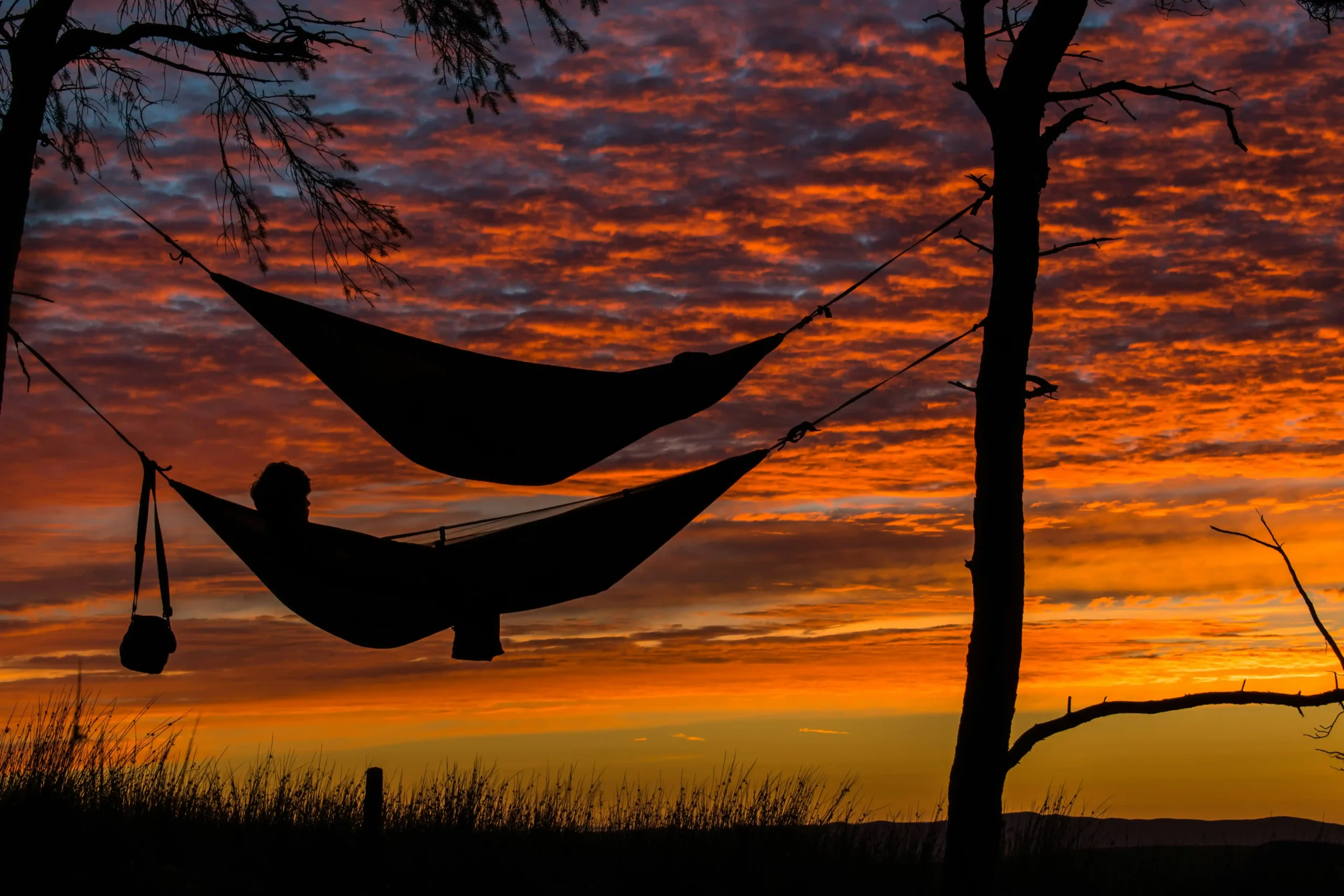 Dos hamacas portátiles colgadas entre árboles al atardecer con siluetas de personas descansando.