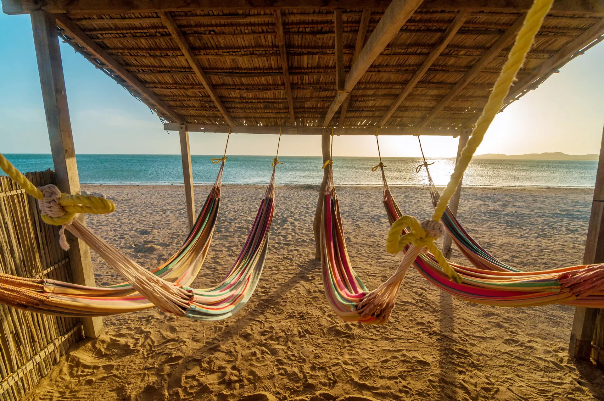 Hamacas de colores bajo un techo de madera frente a una playa de arena dorada al atardecer.