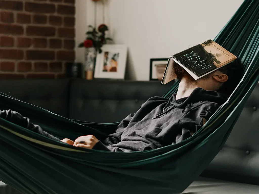 Hombre descansando en una hamaca interior con un libro cubriendo su rostro, en un espacio acogedor con decoración moderna.