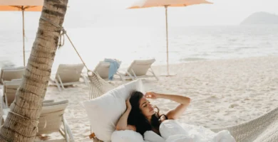 Mujer descansando en una hamaca colgante en la playa, bajo una palmera y junto a sombrillas, disfrutando del sol y la brisa marina.