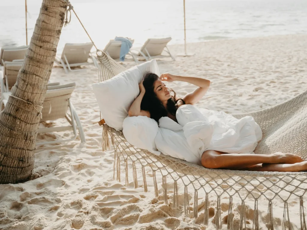 Mujer relajándose en hamaca de red bajo una palmera en la playa, disfrutando del sol y el mar.