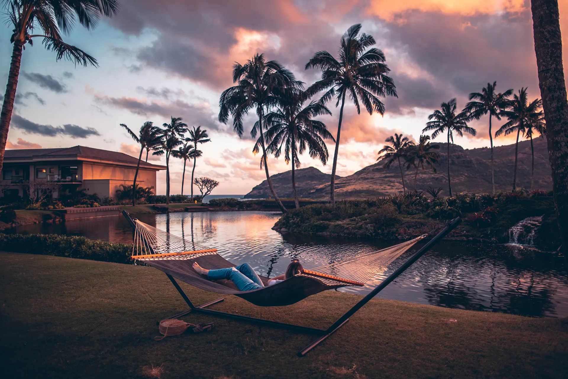 Persona relajándose en una hamaca junto a un lago rodeado de palmeras, con un espectacular atardecer tropical en el horizonte.