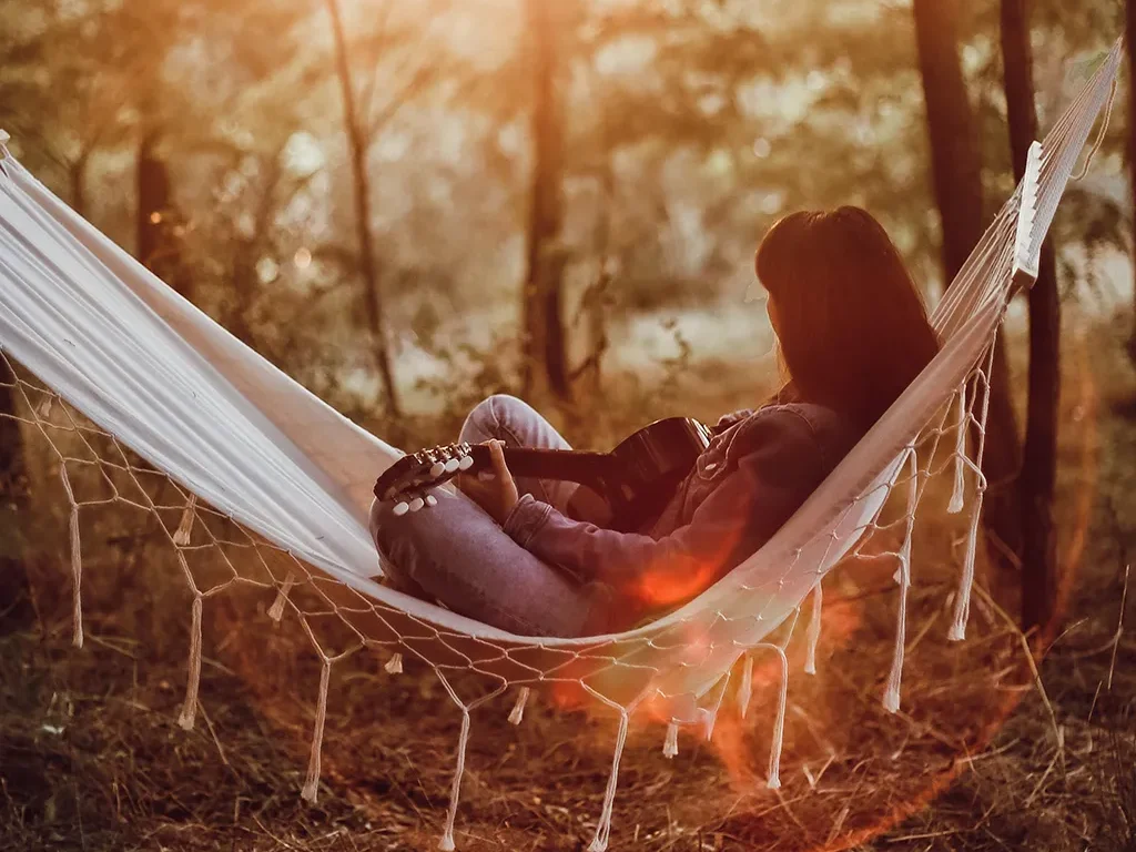 Mujer relajándose en una hamaca blanca en el bosque mientras toca una guitarra al atardecer.