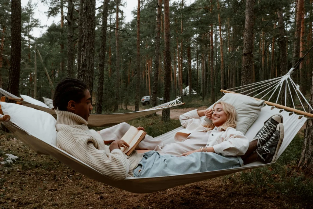 Dos personas disfrutando de una tarde en hamacas colgadas entre árboles en un jardin, uno leyendo un libro y la otra sonriendo relajada