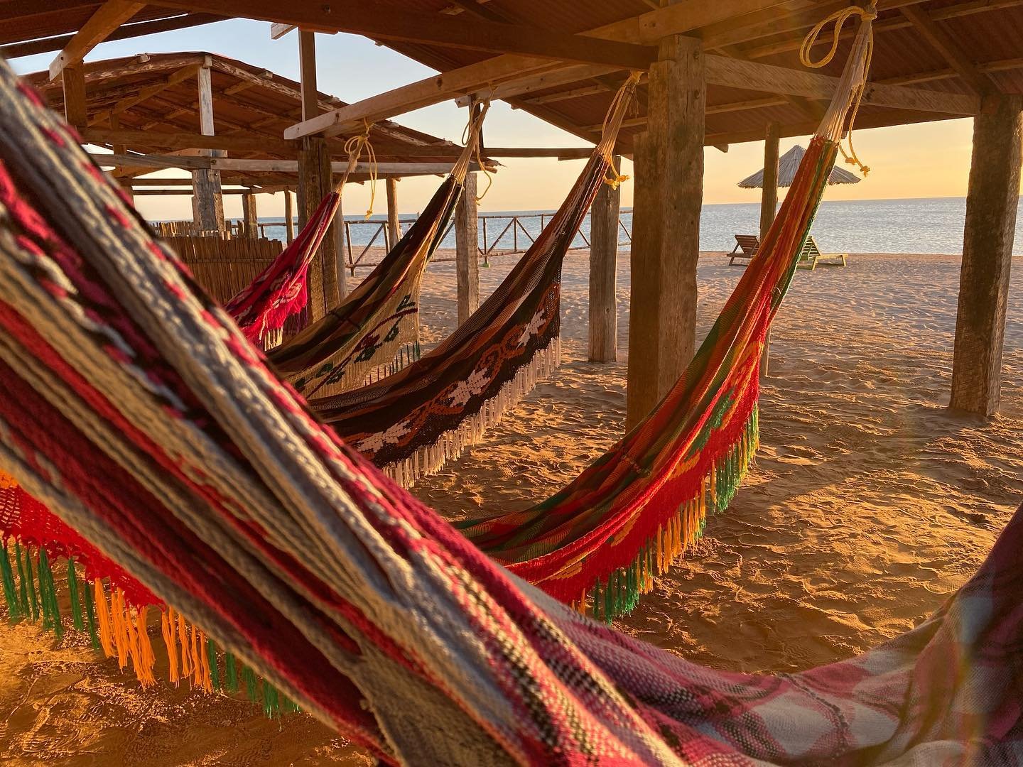 Hamacas de colores colgadas bajo palapas o kioskos de madera en una playa al atardecer.
