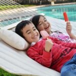Niños sonrientes comiendo paletas en hamaca blanca junto a una piscina.