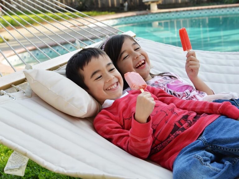 Niños sonrientes comiendo paletas en hamaca blanca junto a una piscina.