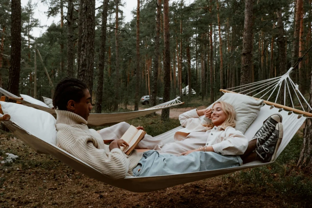 Pareja relajándose en una hamaca de tela blanca en un bosque rodeado de árboles, disfrutando de un momento de lectura y descanso.