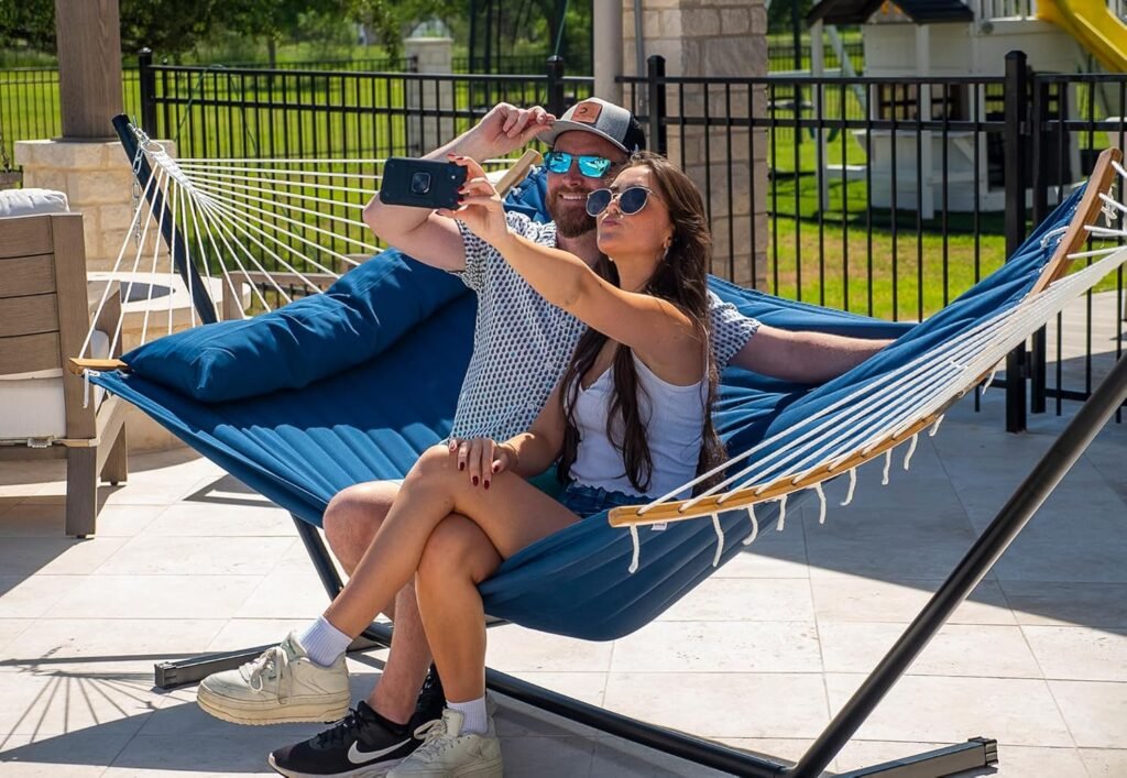 Pareja tomando una selfie mientras se relaja en una hamaca azul acolchada con soporte de acero en terraza exterior.