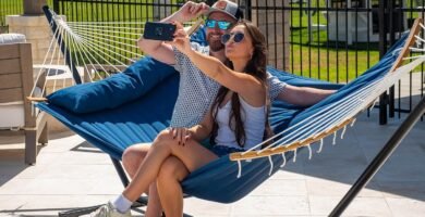 Pareja tomando una selfie mientras se relaja en una hamaca azul acolchada con soporte de acero en terraza exterior.