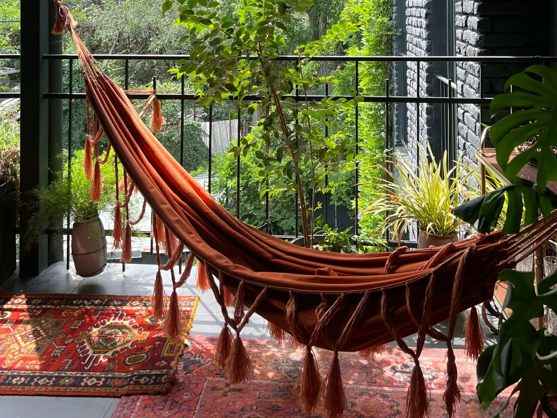 Hamaca de tela color terracota con borlas colgada en balcón rodeado de plantas y alfombras bohemias.