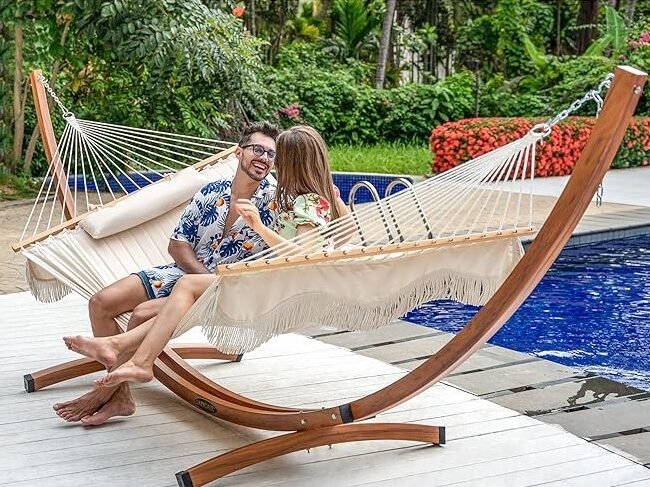 Pareja sentada en una hamaca beige con soporte de madera en una terraza junto a una piscina en un entorno tropical.