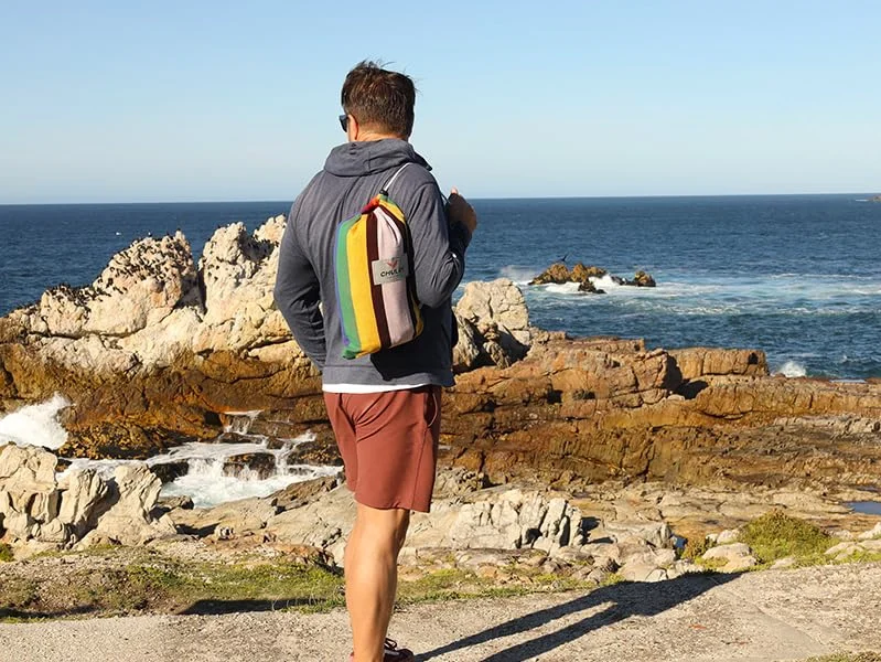 Hombre con mochila de rayas cargando una hamaca portátil mirando al mar desde un acantilado rocoso.