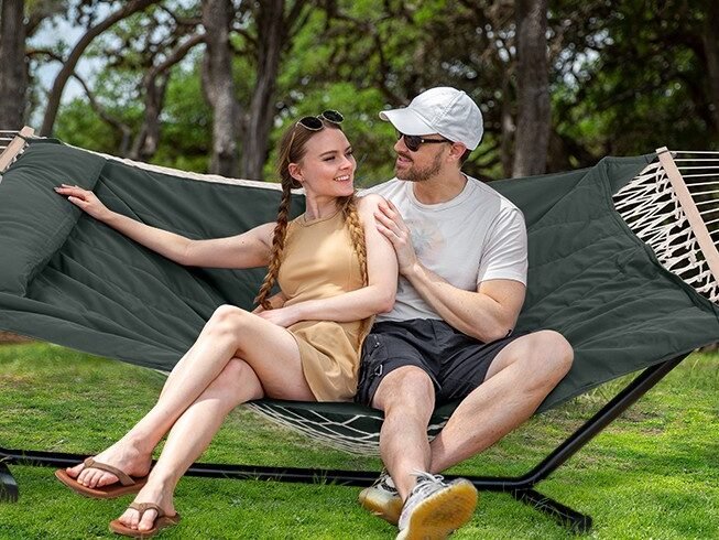 Pareja sentada en una hamaca doble con soporte metálico en un parque arbolado durante un día soleado.