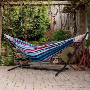 Hamaca de tela de algodón a rayas azules, rojas y blancas con soporte de metal negro ubicada en un jardín rústico con adoquines, cadenas colgantes y vegetación
