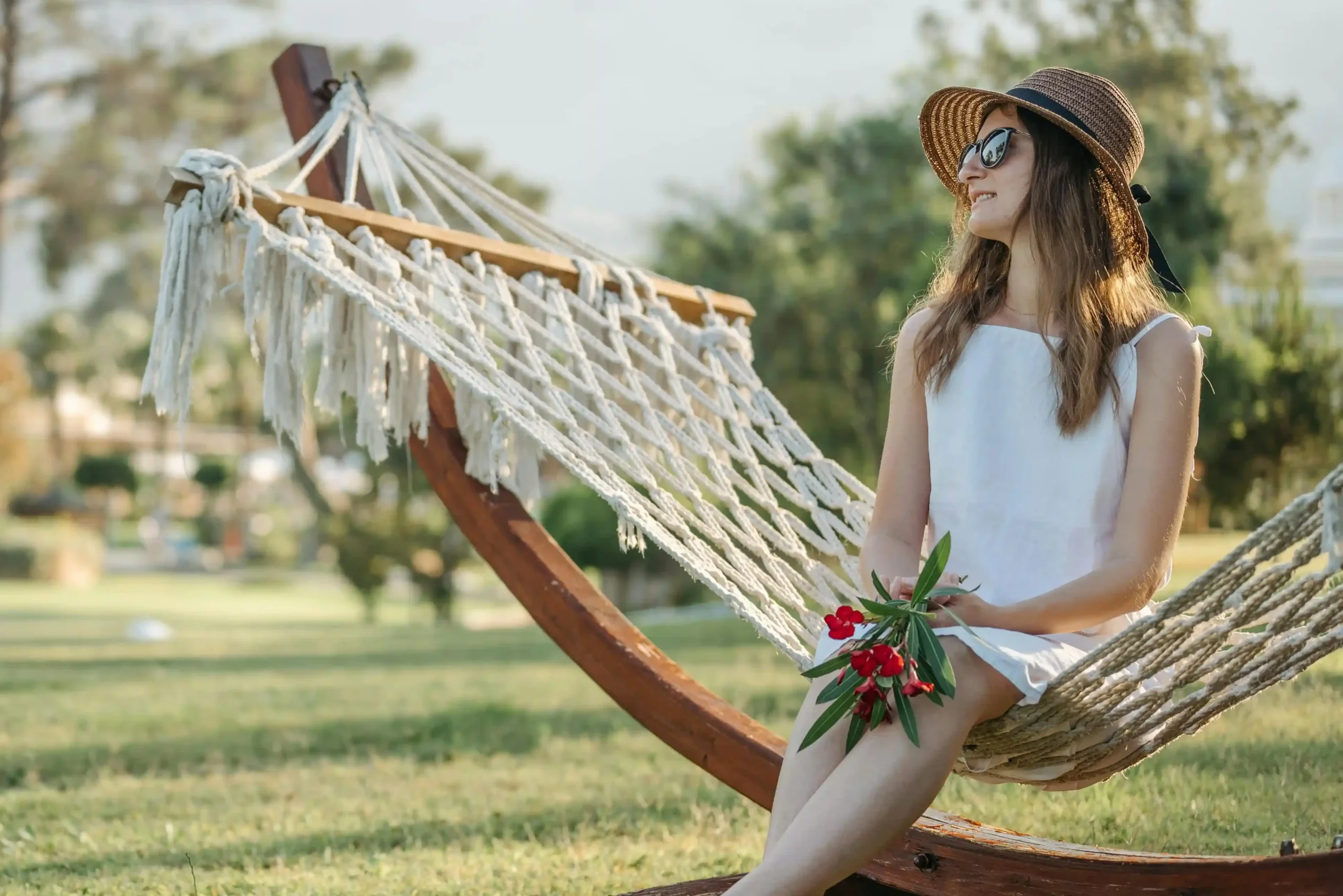 Mujer con gafas de sol y sombrero de ala ancha, sentada en hamaca de cuerda en un jardin con árboles y césped verde
